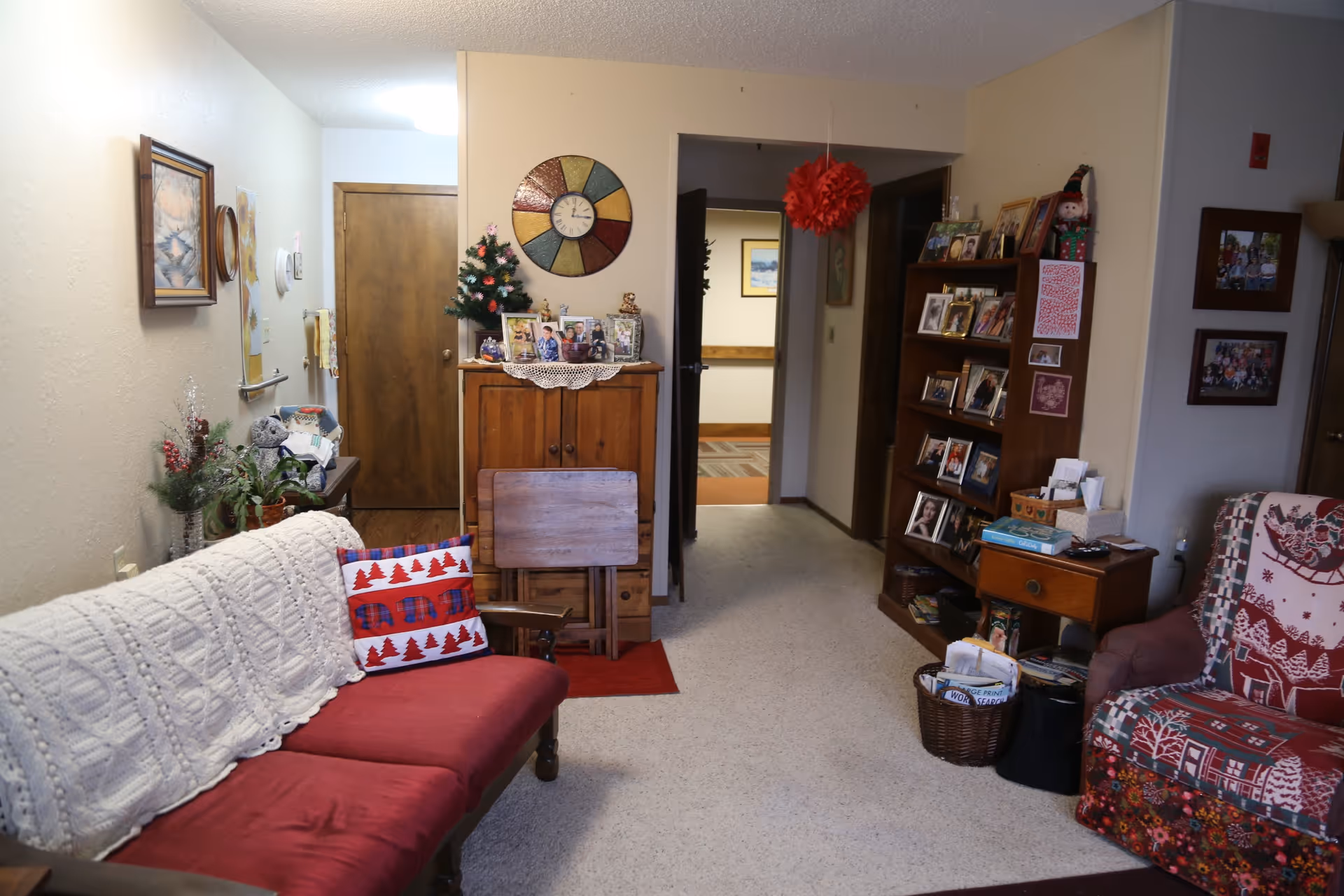 A cozy living room area with a red cushioned wooden sofa covered with a white knitted throw and a decorative pillow with red trees and bears. There is a small wooden cabinet with family photos and a small Christmas tree on top, a colorful wall clock above it, and a wooden bookshelf filled with framed photos and decorations. A red hanging paper decoration is suspended from the ceiling. The room has beige walls, carpeted floor, and wooden doors leading to other rooms.