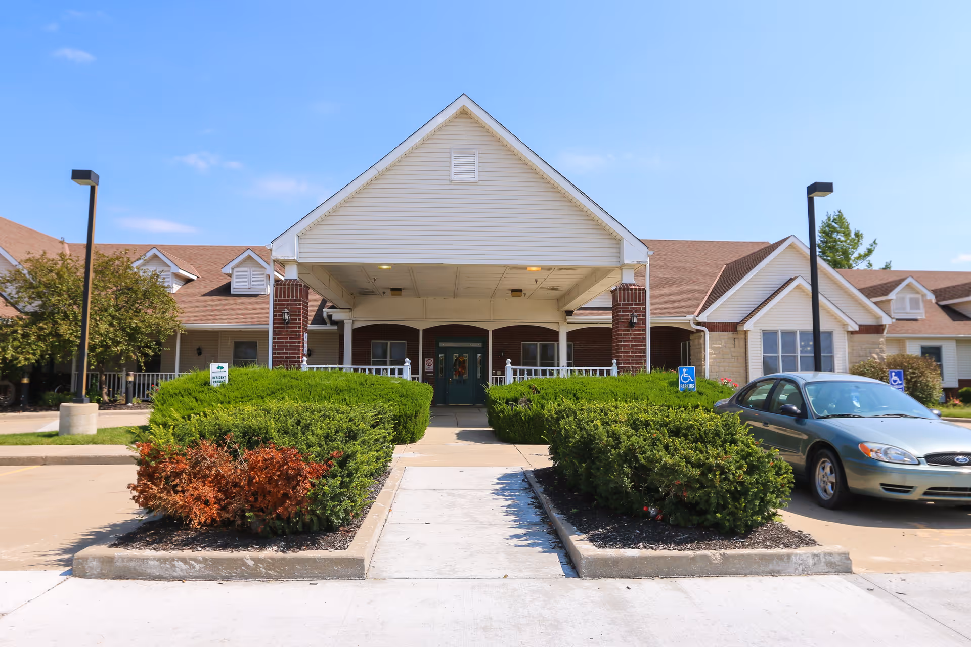Front exterior view of Vintage Park at Gardner, showing a covered entrance with brick pillars, green bushes lining the walkway, a parked car to the right, and a clear blue sky above.