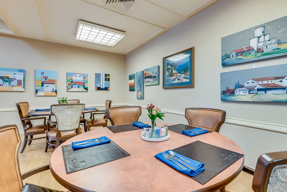Interior view of a dining room with round tables set with blue napkins, silverware, and black placemats. The walls are decorated with various paintings depicting buildings and landscapes. The room has wooden flooring and soft lighting from a ceiling fixture.
