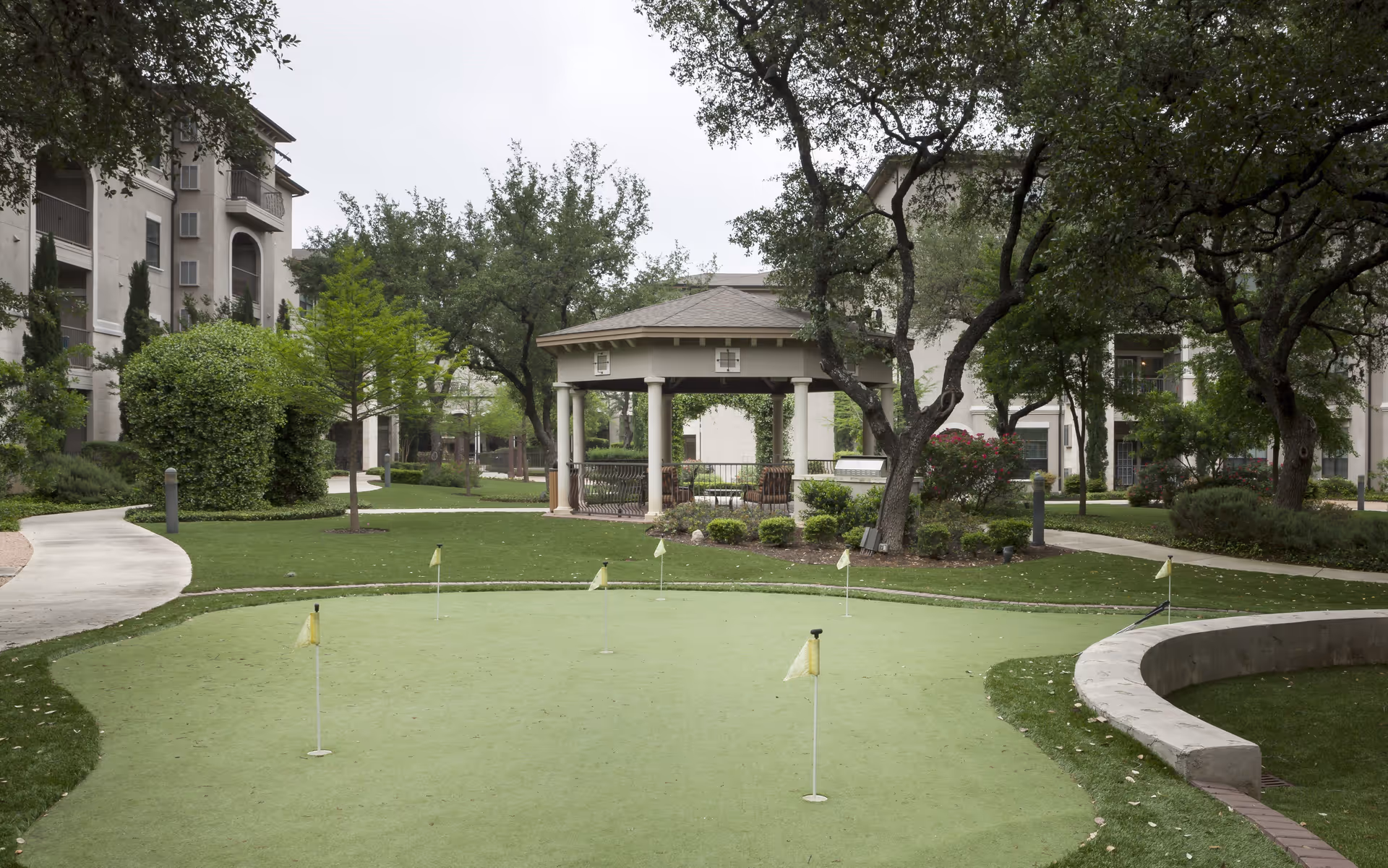 Outdoor putting green with several small flags, surrounded by trees, bushes, and a curved concrete bench. In the background, there is a gazebo with seating and multi-story residential buildings.