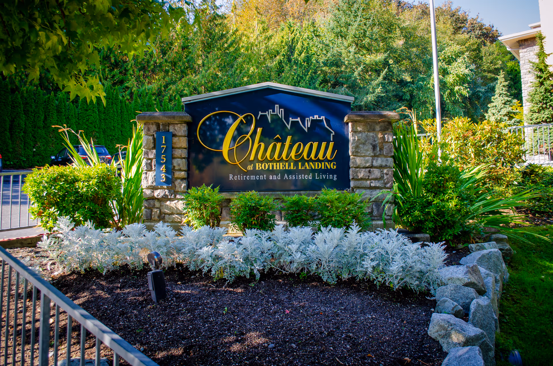Stone sign for Chateau at Bothell Landing Retirement and Assisted Living surrounded by green shrubs and plants, with a background of tall green trees and a clear blue sky.