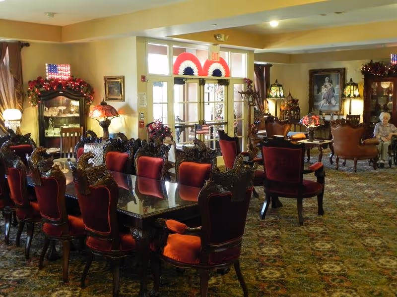 Ornate dining table with red upholstered chairs in a decorated senior living common area with lamps and additional seating.