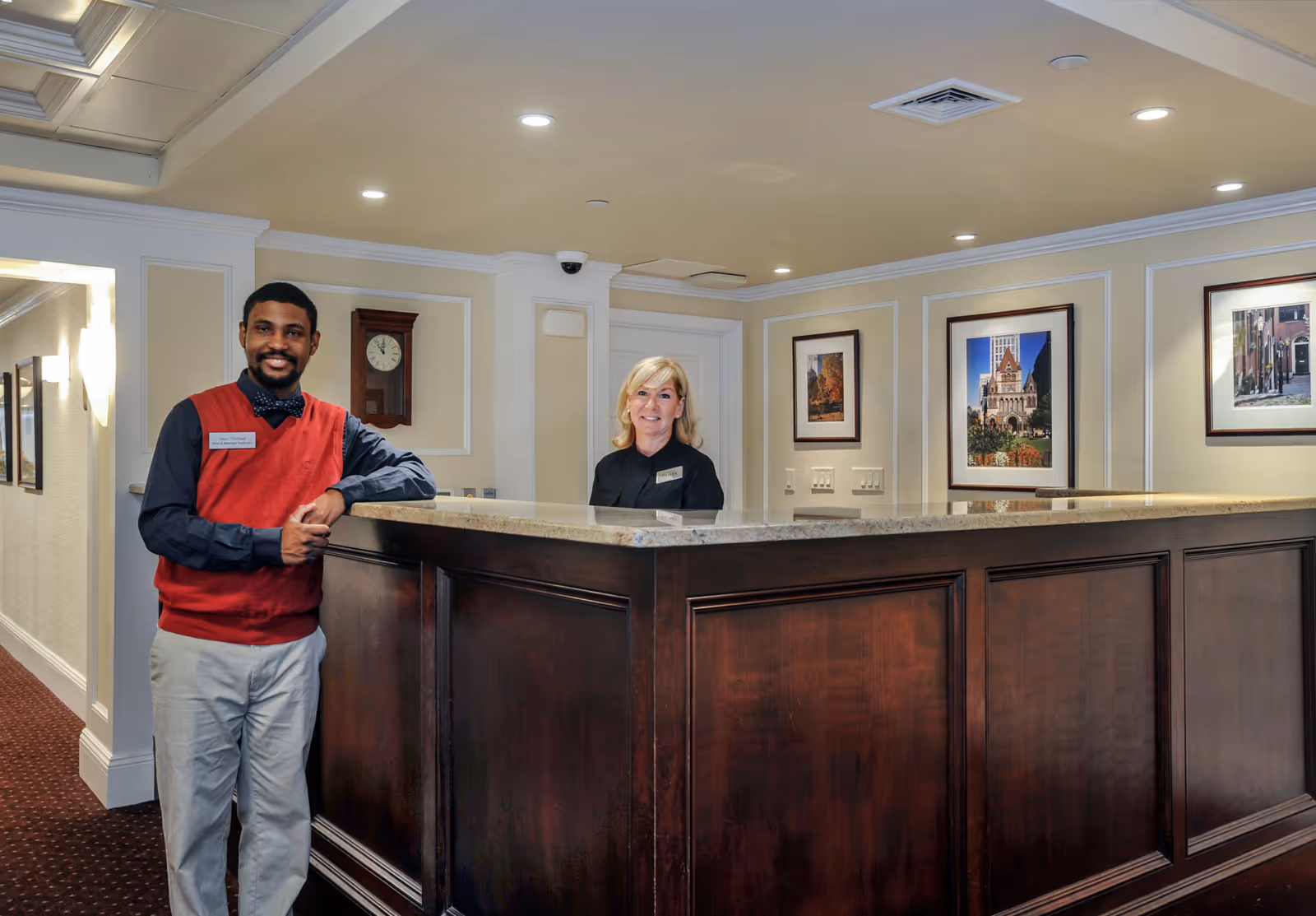 A man and a woman standing behind a dark wooden reception desk in a well-lit interior space. The man is wearing a red vest over a dark shirt and light pants, smiling with his hands resting on the desk. The woman is behind the desk, also smiling, with framed pictures on the wall behind her and a clock mounted on the wall to the left.