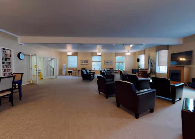 Spacious communal lounge with multiple armchairs, tables, windows, and a TV/fireplace.