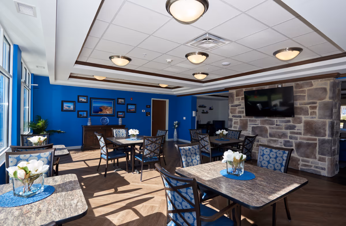 Bright communal dining area with several tables and chairs, blue accent walls, a stone TV wall, and large windows.