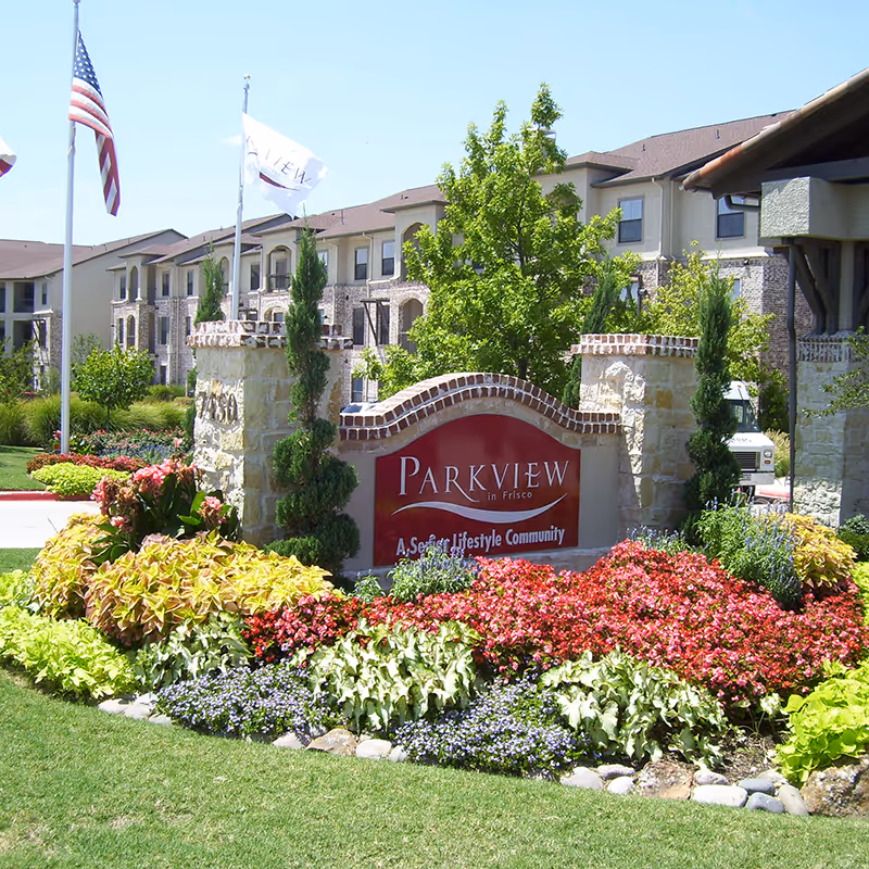Entrance sign for Parkview in Frisco, a senior lifestyle community, surrounded by colorful flowers and greenery with a multi-story residential building in the background and two flagpoles displaying the American flag and another flag.