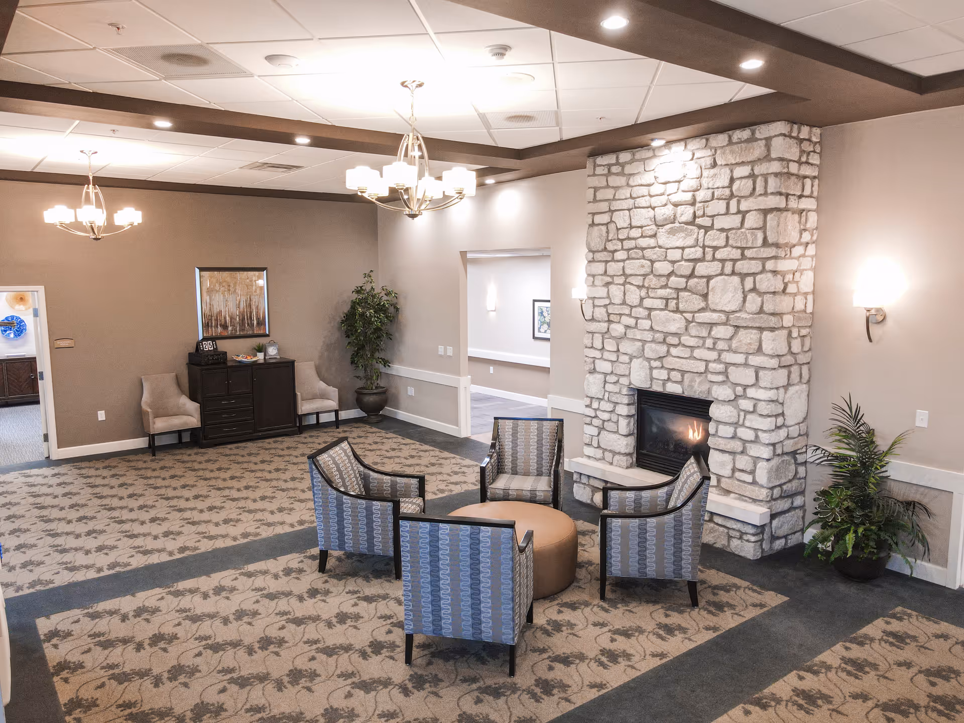 A cozy senior living facility common area with a stone fireplace, four patterned armchairs arranged around a round ottoman, beige walls, carpeted floor with a floral pattern, two chandeliers, and potted plants near the walls.