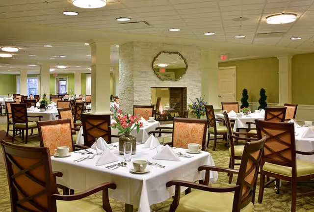 Bright dining room with tables set in white linens, wooden chairs, floral centerpieces, and a central fireplace.