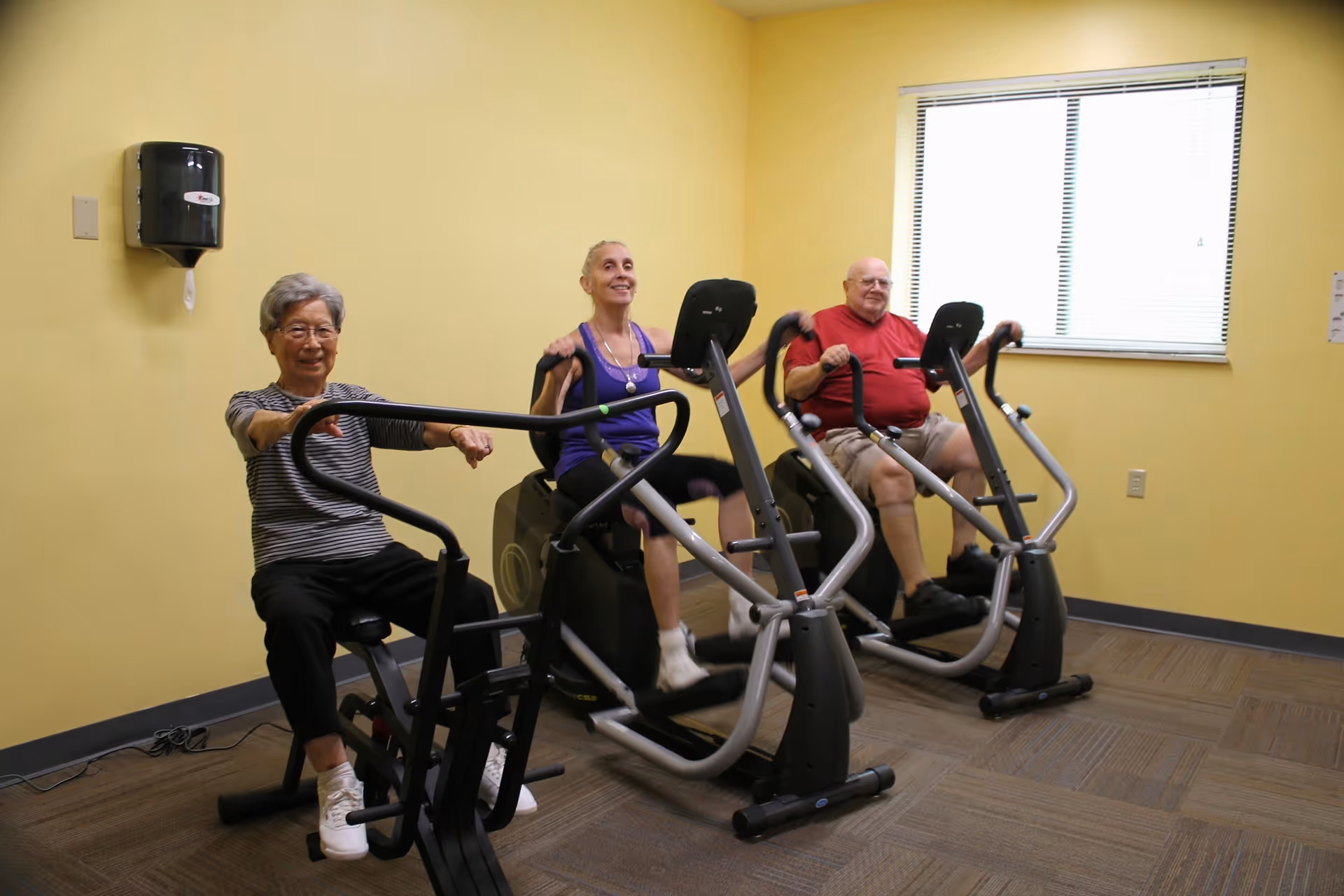 Three elderly individuals exercising on stationary elliptical machines in a bright room with yellow walls and a window with blinds.