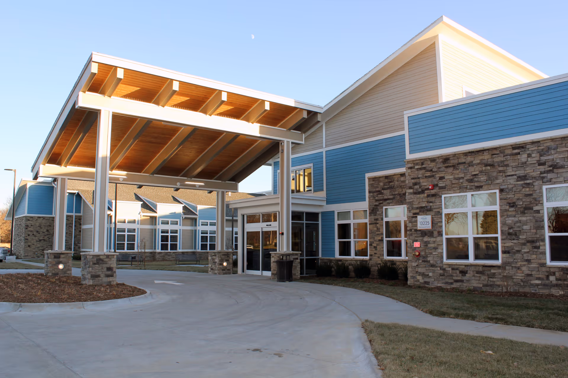Exterior view of Hillcrest Millard facility showing a covered entrance with stone pillars and a driveway leading up to the main doors. The building features blue siding with stone accents and multiple windows under a clear sky.