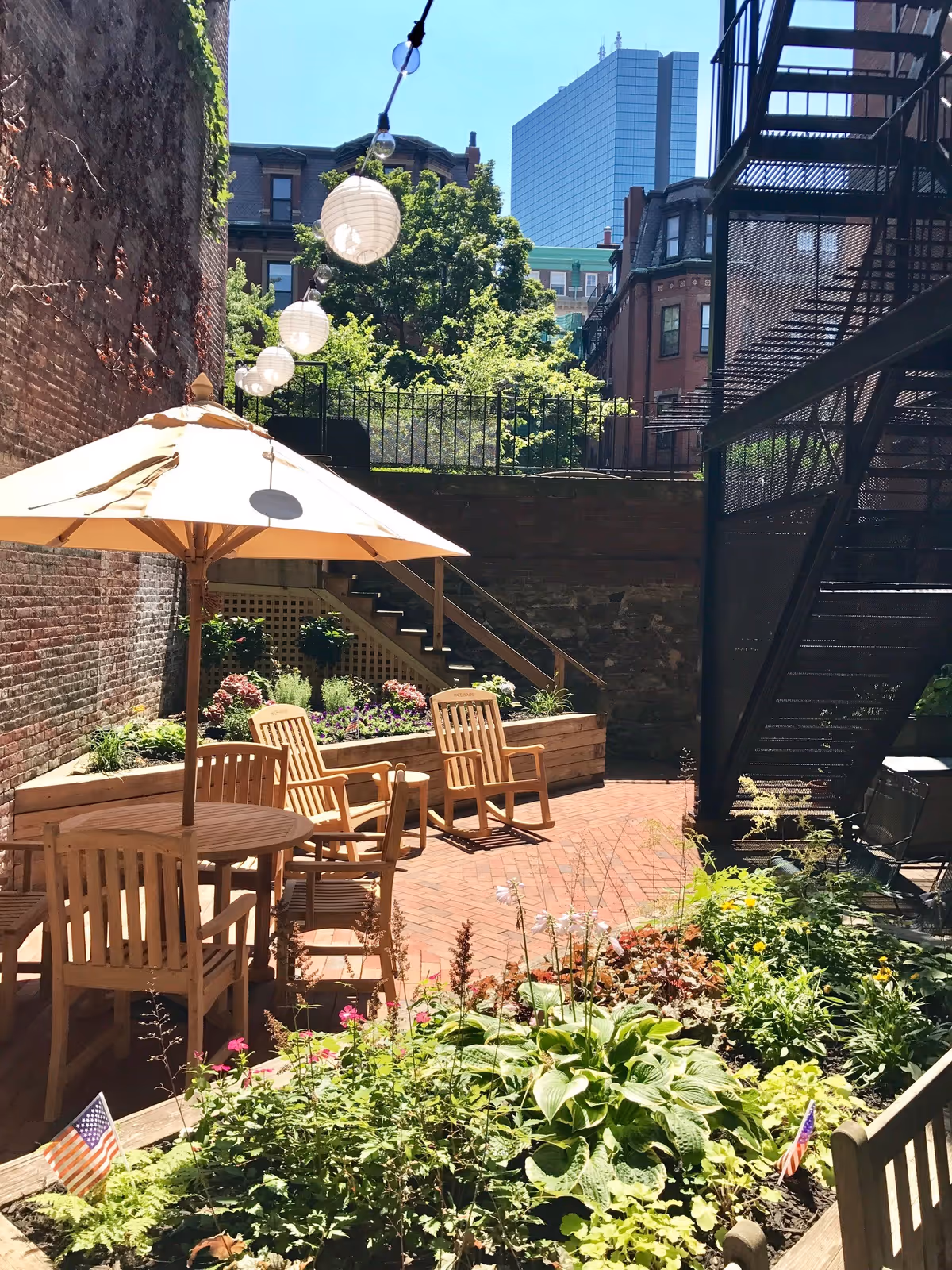 Outdoor patio area with wooden tables and chairs under a large beige umbrella, surrounded by flower beds and greenery. String lights with round paper lanterns hang above. Brick walls and stairs are visible, along with a fire escape on the right side. Tall buildings and trees are in the background under a clear blue sky.