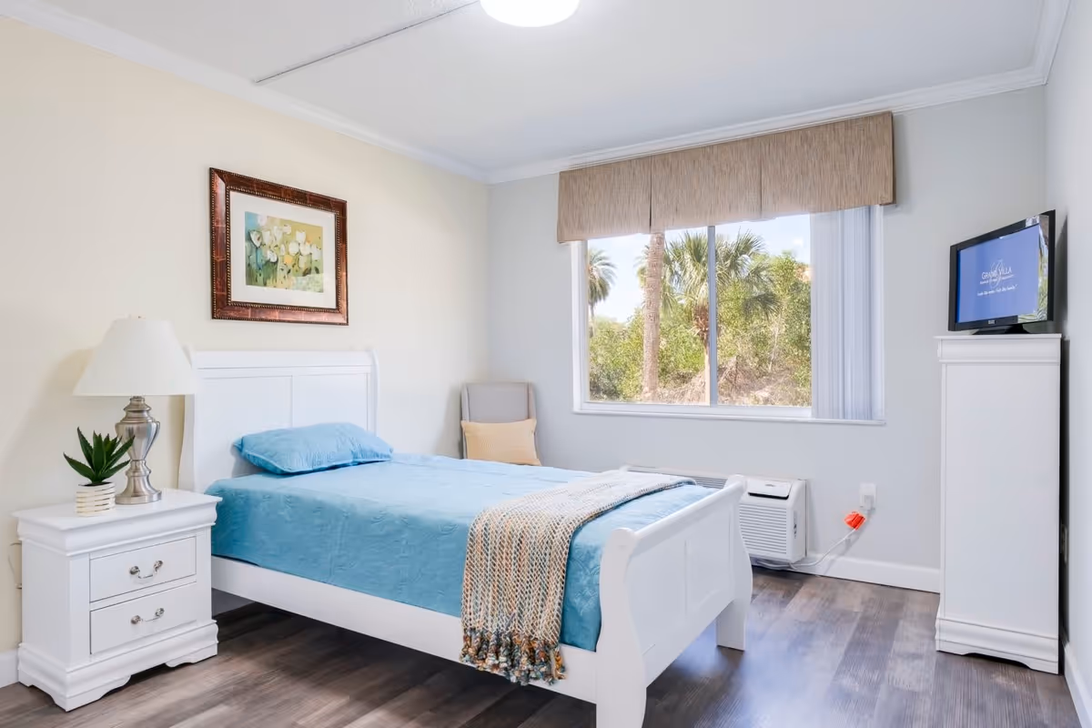 A bright and clean bedroom with a white wooden bed frame and a blue bedspread. There is a white nightstand with a silver lamp and a small potted plant. A framed floral painting hangs on the wall above the bed. A window with beige valance shows palm trees outside. A white dresser with a TV on top is positioned near the window. The room has wood flooring and light-colored walls.