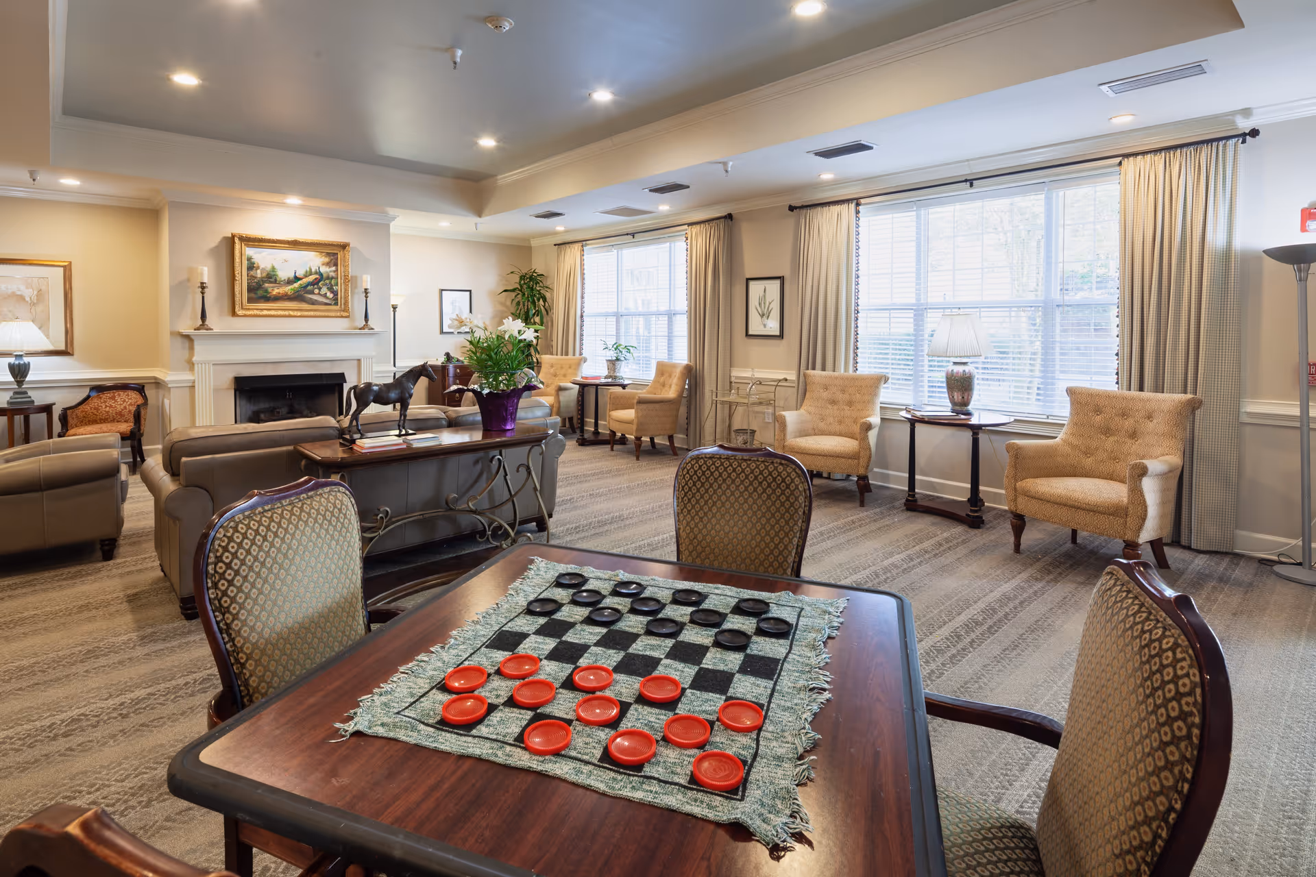 A cozy living room area in a senior living facility with a checkers game set up on a wooden table in the foreground. The room features several upholstered armchairs and sofas arranged around a fireplace with a painting above it. Large windows with curtains allow natural light to fill the space, and there are side tables with lamps and decorative plants throughout the room.