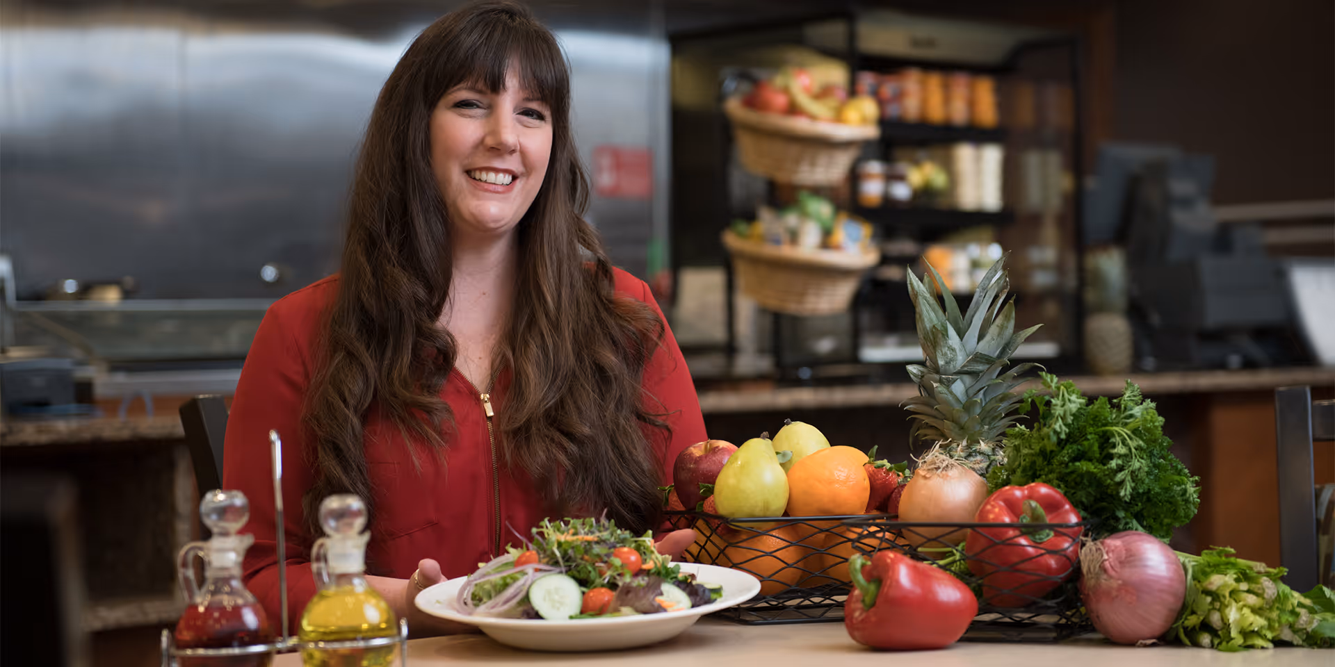 A woman with long brown hair wearing a red top is sitting at a table in a kitchen or dining area. In front of her is a plate of fresh salad and a basket filled with various fruits and vegetables including pears, oranges, pineapple, onions, bell peppers, and leafy greens. The background shows kitchen equipment and shelves with more produce.