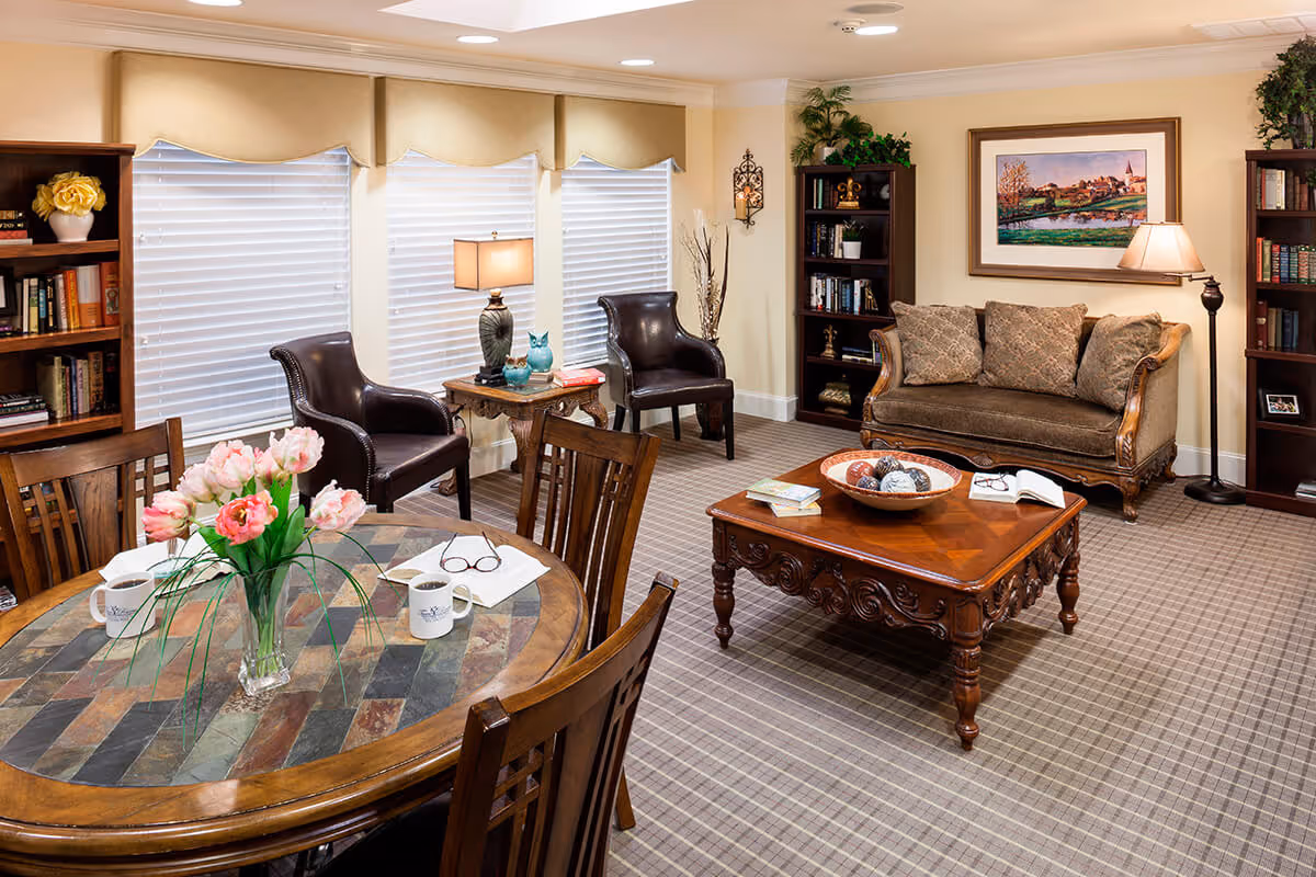 A cozy senior living room with a patterned carpet, a wooden coffee table with decorative items, a cushioned sofa with three pillows, two dark leather armchairs, a round wooden dining table with four chairs, a vase of pink flowers, two coffee mugs, and reading glasses. The room has bookshelves filled with books, a floor lamp, a table lamp, and a framed landscape painting on the wall.