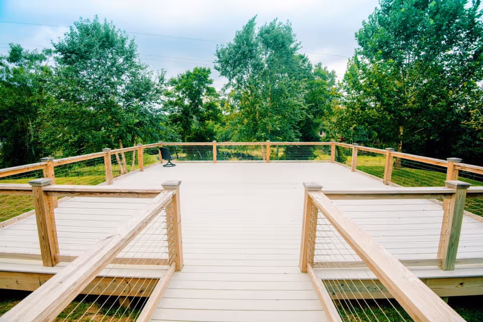 A spacious wooden outdoor deck with railings and a single black chair, surrounded by green trees and grass under a cloudy sky.