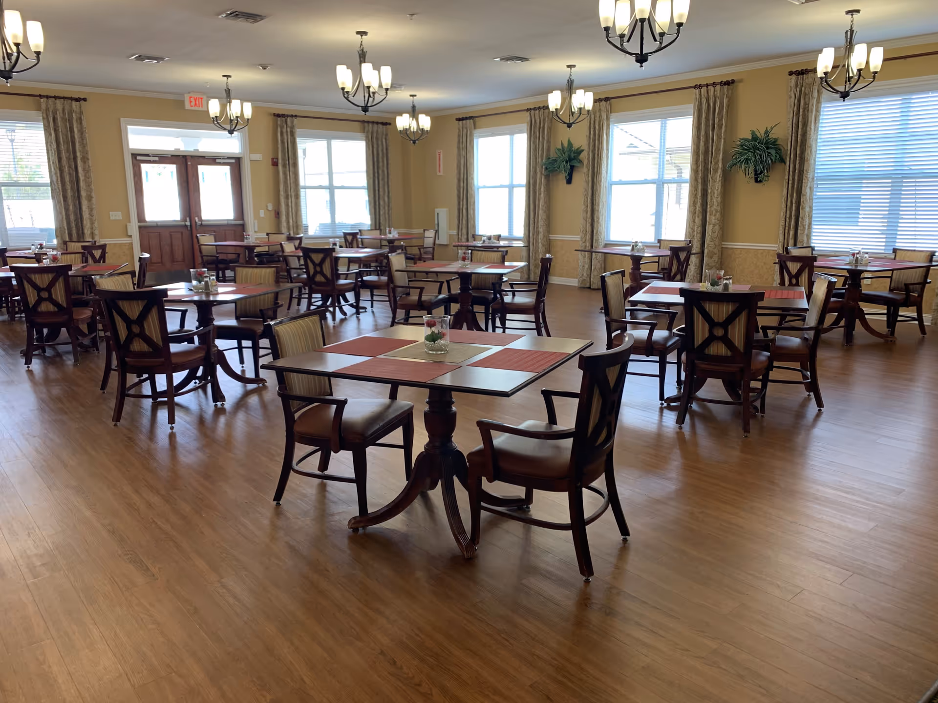 A spacious dining room with multiple wooden tables and chairs arranged neatly. Each table has placemats and a small decorative centerpiece. The room has large windows with curtains, wooden flooring, and several ceiling light fixtures providing warm lighting.
