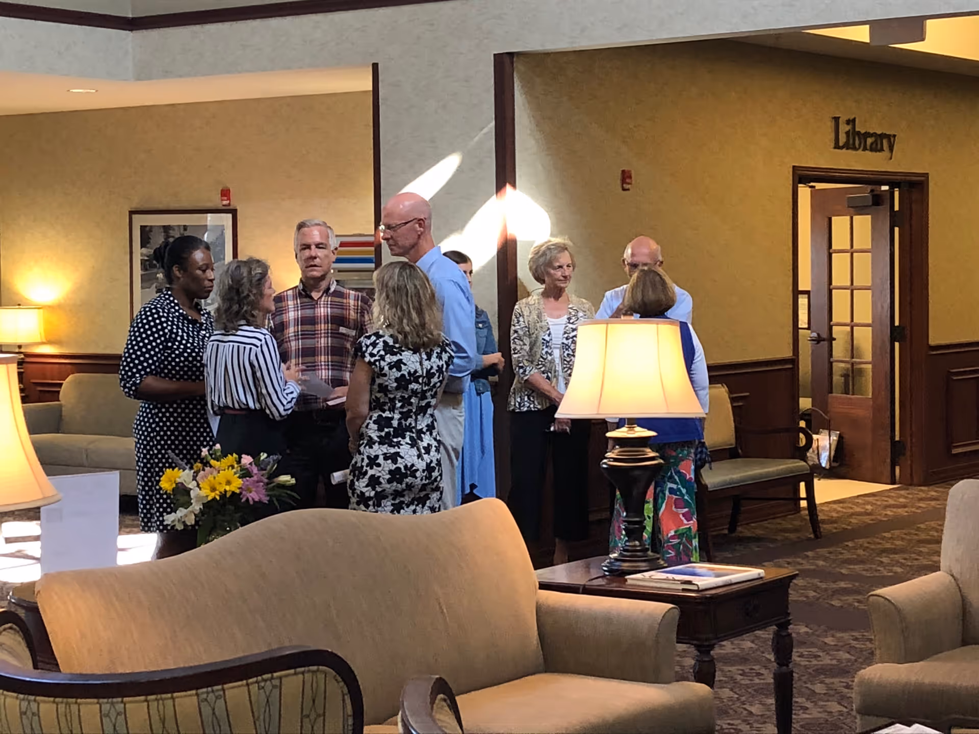 A group of seven people standing and conversing in a well-lit common area with comfortable seating, lamps, and a sign indicating the entrance to a library.