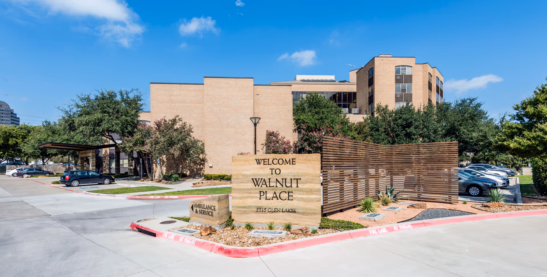 Exterior view of Walnut Place senior living facility with a large stone sign reading 'Welcome to Walnut Place 5515 Glen Lakes' in front. The building is a multi-story brick structure surrounded by trees and a parking lot with several cars. The sky is clear and blue.