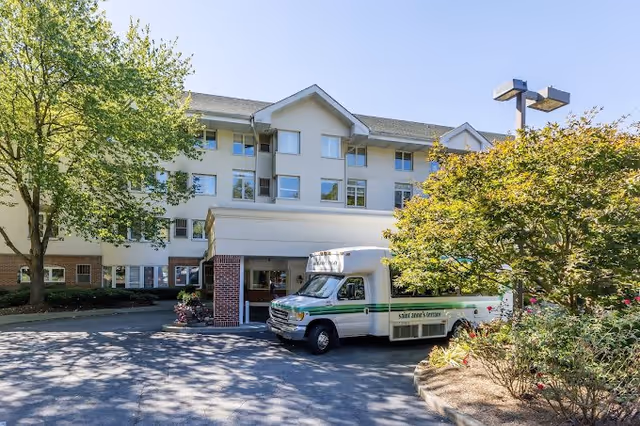Exterior view of a multi-story senior living facility named Saint Anne's Terrace with a white shuttle bus parked in front. The building is surrounded by trees and landscaping under a clear blue sky.