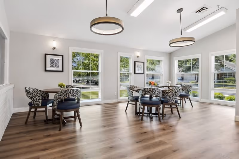 A bright dining room with large windows letting in natural light. The room features wooden flooring, two round tables each surrounded by four patterned upholstered chairs with dark blue seats. There are two modern circular pendant lights hanging from the ceiling and framed artwork on the light gray walls.
