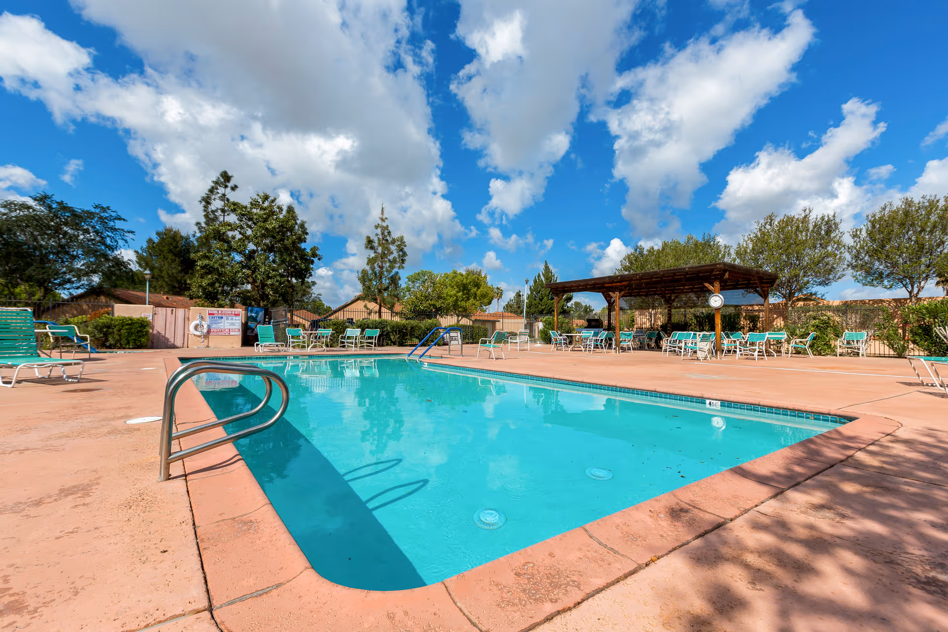 Outdoor swimming pool and deck with lounge chairs and a shaded seating pavilion under a blue sky.