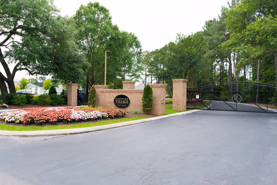 Entrance gate to Plantation Village featuring a brick wall with a sign, black metal gate, and surrounding greenery including trees and flower beds.