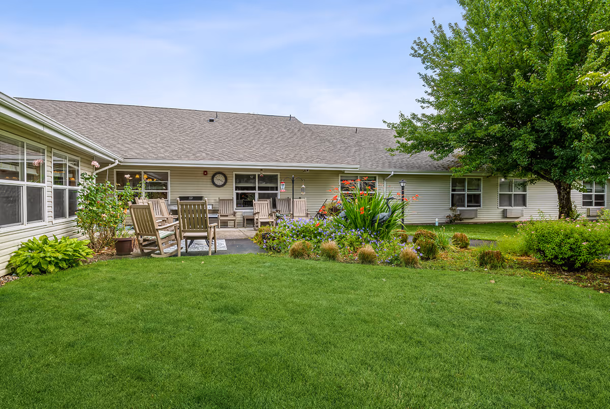 Outdoor courtyard area of a senior living facility with green grass, garden beds with flowers and shrubs, several wooden chairs arranged on a paved patio, and a large tree on the right side. The building has beige siding and multiple windows.