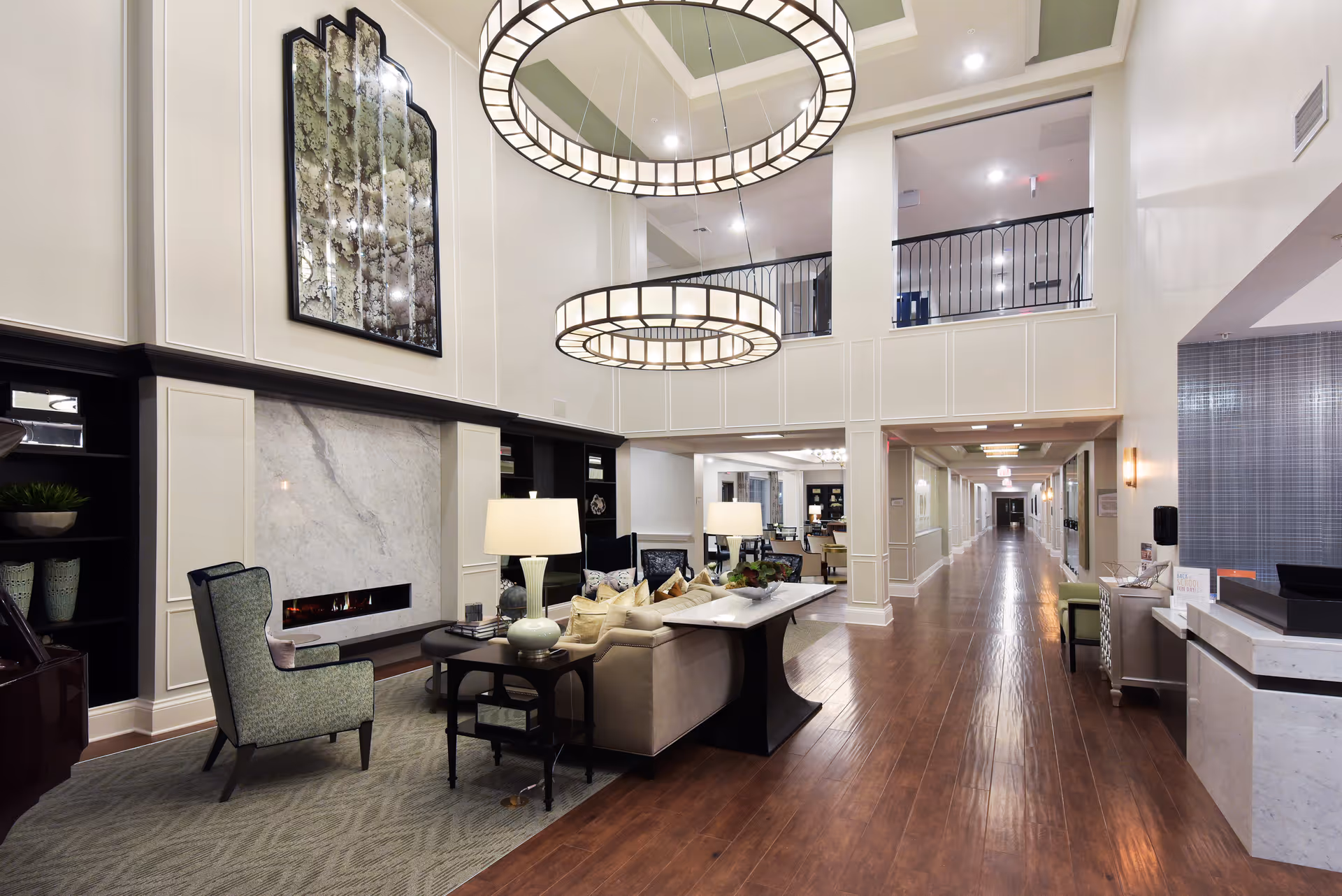A spacious and elegant senior living facility common area with high ceilings, two large circular chandeliers, a fireplace with a marble surround, comfortable seating including armchairs and a sofa, decorative lamps, and a long hallway leading to other rooms.