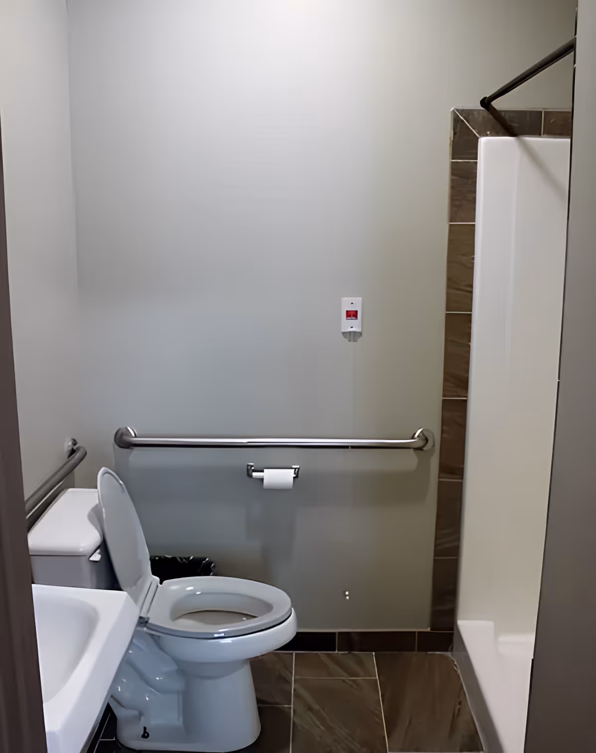 Small accessible bathroom with a toilet (seat up), wall grab bars, a shower stall, sink, and tiled floor.