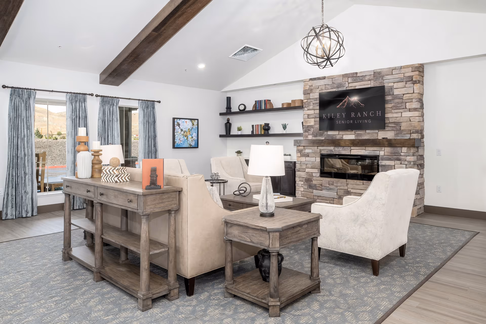 A bright and spacious living room in a senior living facility featuring beige upholstered chairs arranged around a wooden coffee table and side table with a lamp. The room has a stone fireplace with a mounted TV displaying the Kiley Ranch Senior Living logo. There are shelves with decorative items on the wall and large windows with blue curtains letting in natural light. The ceiling has exposed wooden beams and a modern chandelier.