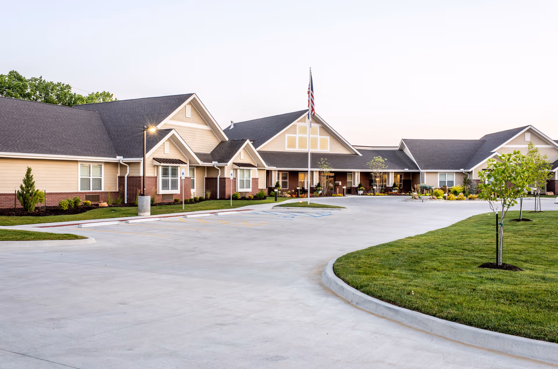 Exterior view of Cross Creek at Lee's Summit Memory Care & Assisted Living facility showing a single-story building with beige siding and brick accents, a large paved driveway with parking spaces, a flagpole with an American flag, and landscaped green areas with small trees and shrubs.
