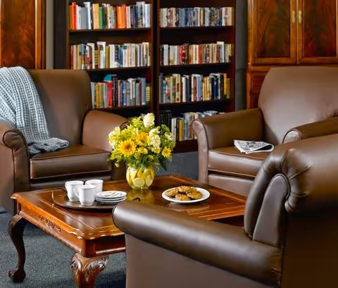 A cozy living room area with three brown leather armchairs arranged around a wooden coffee table. The table holds a vase with yellow and white flowers, a plate of cookies, and a tray with cups and saucers. In the background, there is a bookshelf filled with books and a wooden cabinet.