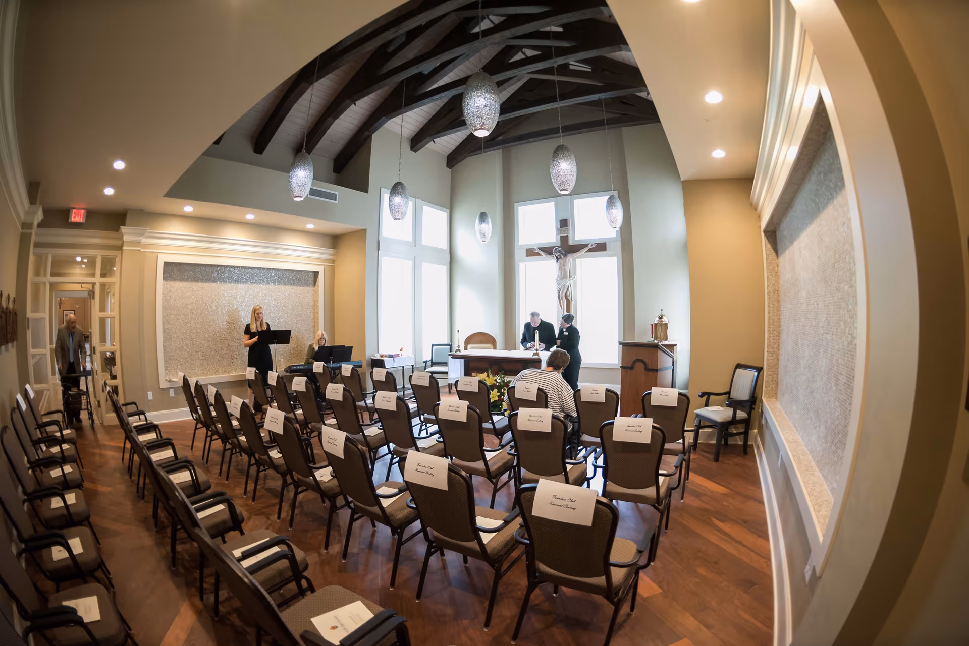 Interior of a chapel or small worship room with rows of chairs facing an altar. A large crucifix is mounted on the wall behind the altar. Several people are present, including a woman standing and reading from a music stand, a person playing a keyboard, and two people near the altar. The room has high ceilings with exposed wooden beams and hanging pendant lights.