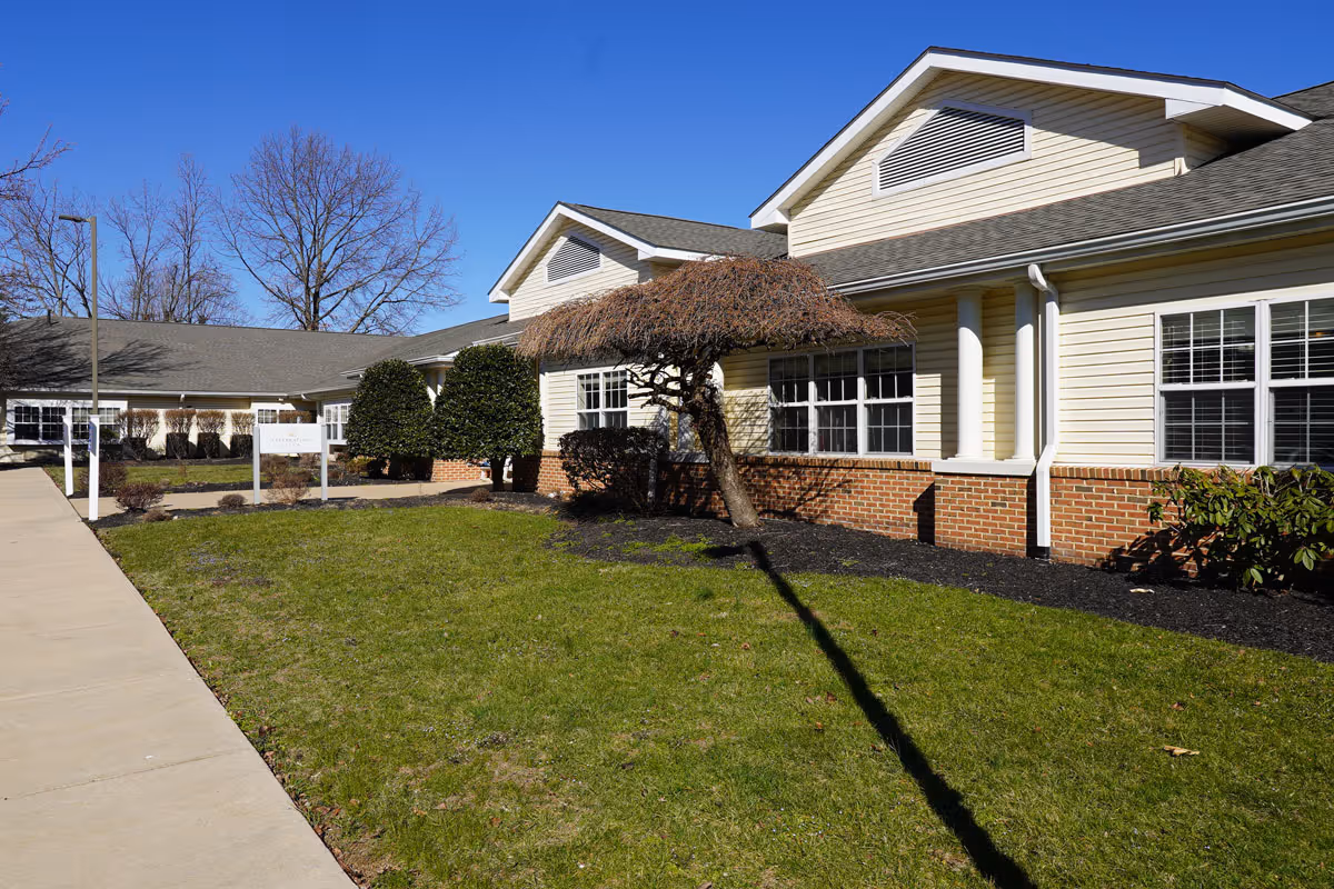 Exterior view of Celebration Villa of Martinsburg, showing a single-story building with beige siding and brick accents, a manicured lawn, trimmed bushes, and a small tree with bare branches under a clear blue sky.