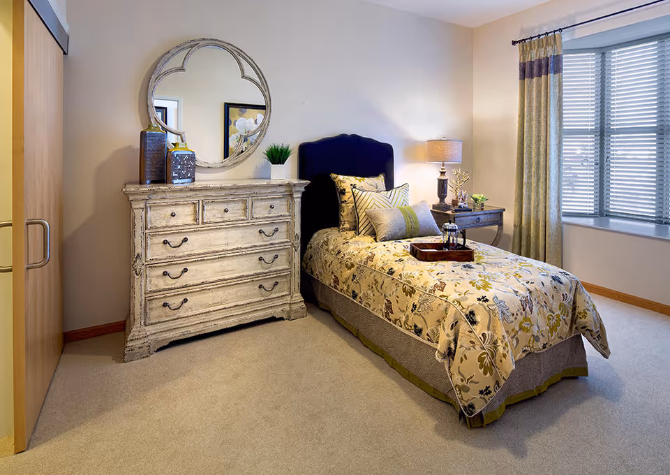 A cozy bedroom with a single bed covered in a floral patterned bedspread and multiple pillows. Next to the bed is a wooden nightstand with a lamp and decorative items. Across from the bed is a distressed white dresser with drawers and a round mirror hanging above it. A window with blinds and curtains allows natural light into the room.