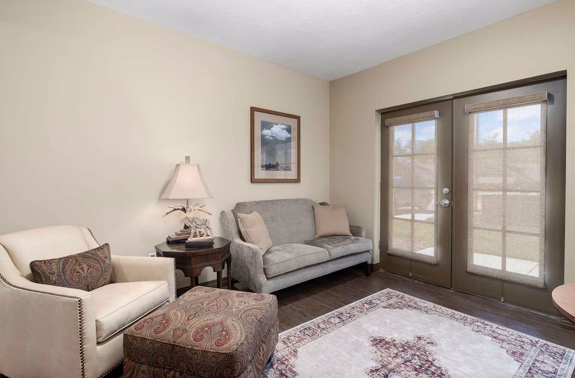 Cozy living room with an armchair and ottoman, a sofa, side table with lamp, framed art, rug, and French doors with shades.