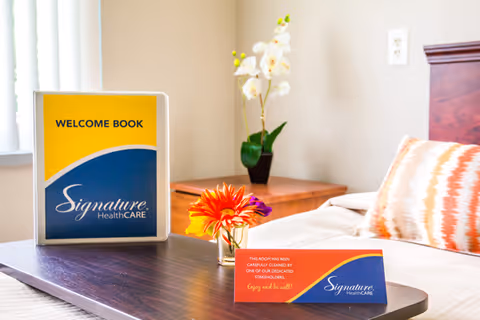 A neatly made bed with a patterned pillow in a room at Signature HealthCARE of Clarksville. On a dark wooden tray placed on the bed, there is a welcome book with the Signature HealthCARE logo, a small vase with colorful flowers, and a card indicating the room has been carefully cleaned. A wooden nightstand with a white orchid plant is visible in the background.