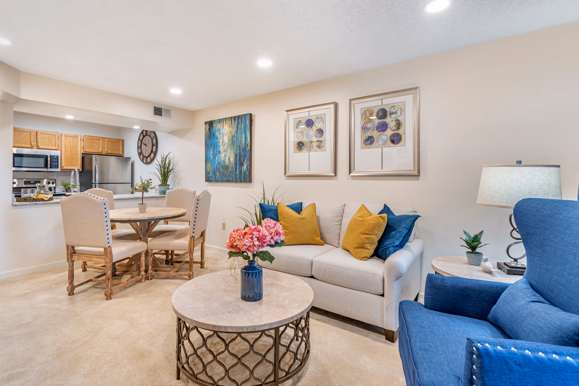 A cozy living room and dining area with beige carpet and walls. The living room features a beige sofa with blue and yellow throw pillows, a blue armchair, a round marble coffee table with a blue vase holding pink flowers, and a side table with a lamp and small plant. The dining area has a round wooden table with four beige upholstered chairs. The kitchen is visible in the background with wooden cabinets, stainless steel appliances, and a large wall clock. The walls are decorated with framed abstract artwork.