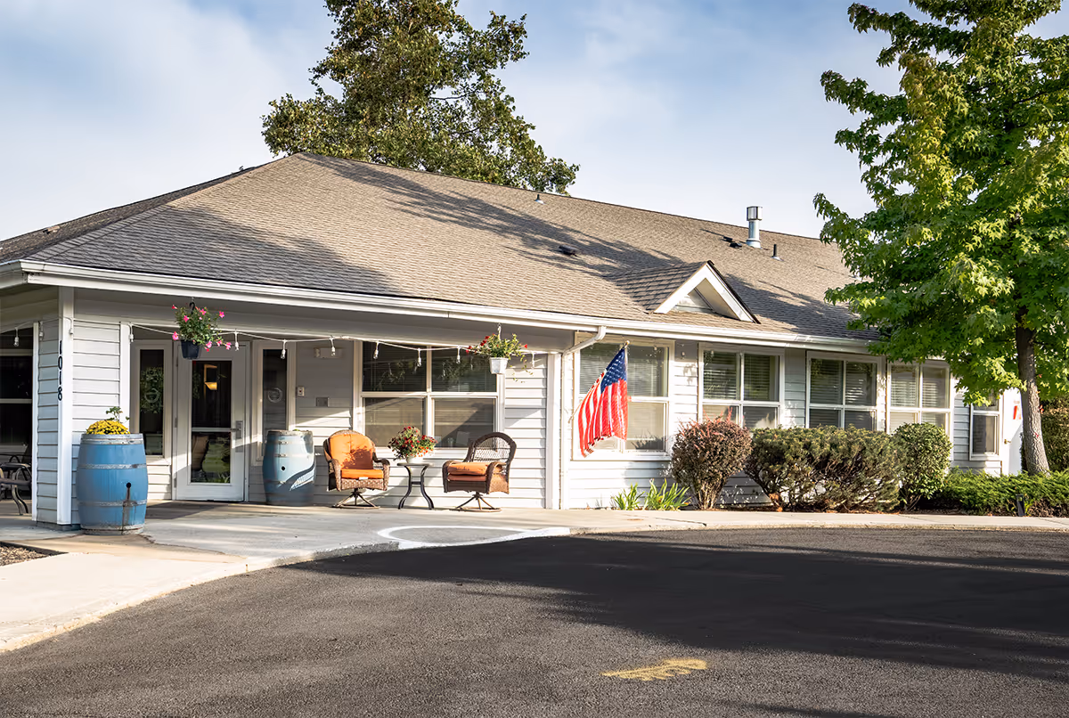 Exterior view of a single-story assisted living facility building with white siding and a gray shingled roof. There are two orange cushioned chairs and a small table on the front porch, along with two blue barrels and hanging flower pots. An American flag is displayed near the windows, and there are bushes and a tree around the building. The driveway and sidewalk are visible in the foreground.