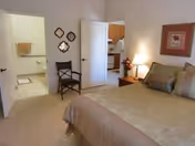 A cozy bedroom with a large bed, a chair, and decorative wall art. A doorway leads to a bathroom in the background, and a glimpse of a kitchen can be seen through another door.
