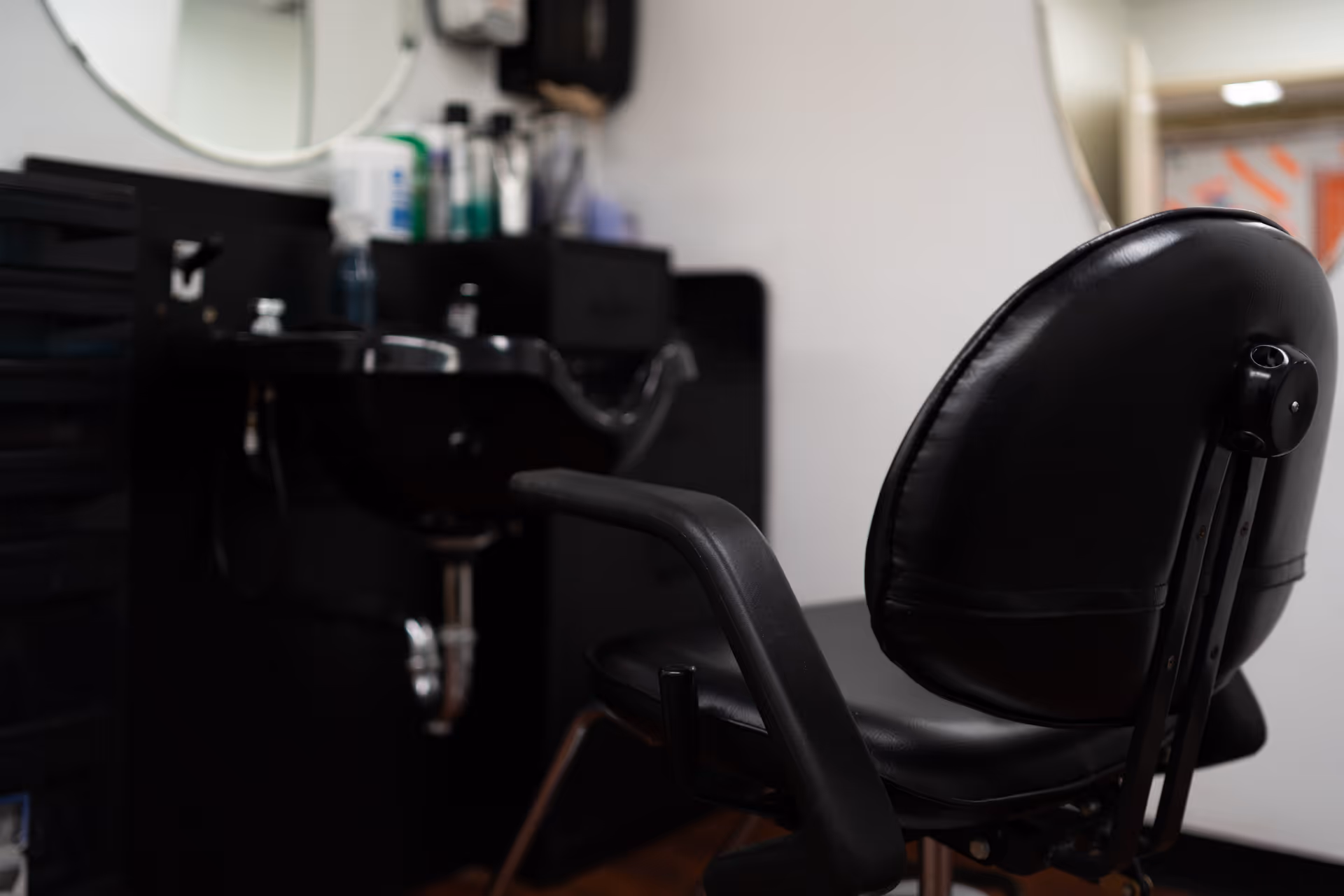 A close-up view of a black salon chair in front of a black hair washing sink in a hair salon or barber area. Various hair care products and tools are visible on the counter behind the sink, and a round mirror is partially visible on the wall.