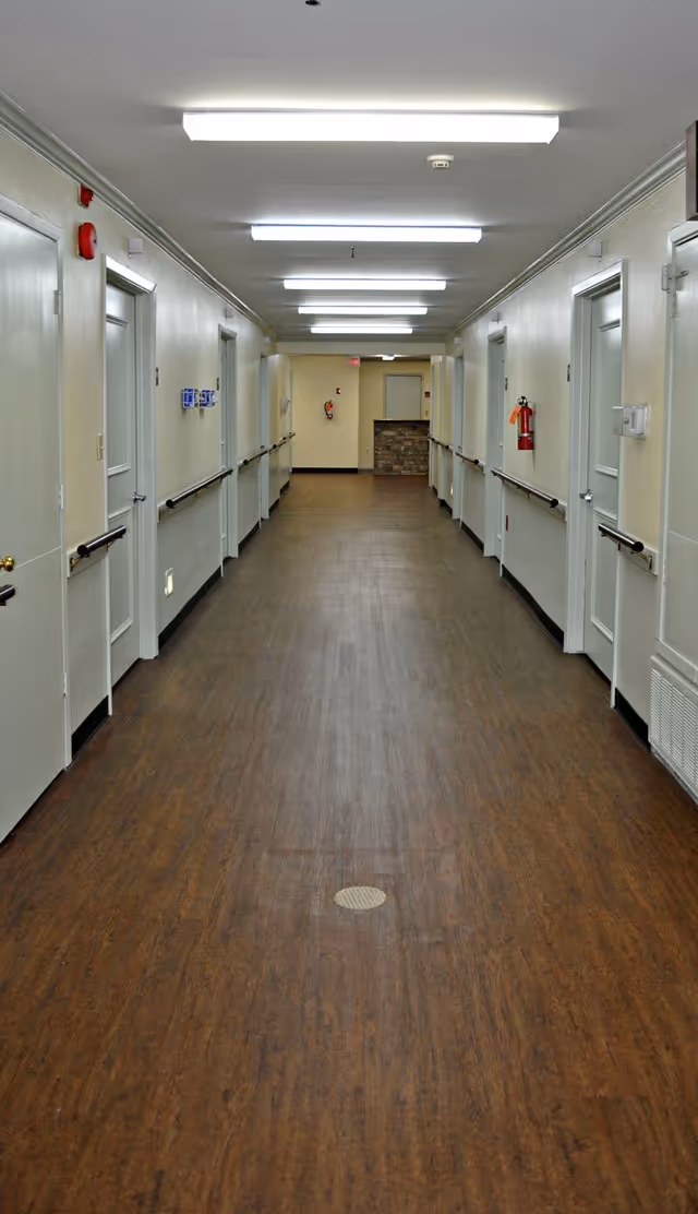 Well-lit long nursing home corridor with doors and handrails on both sides and wood-look flooring.