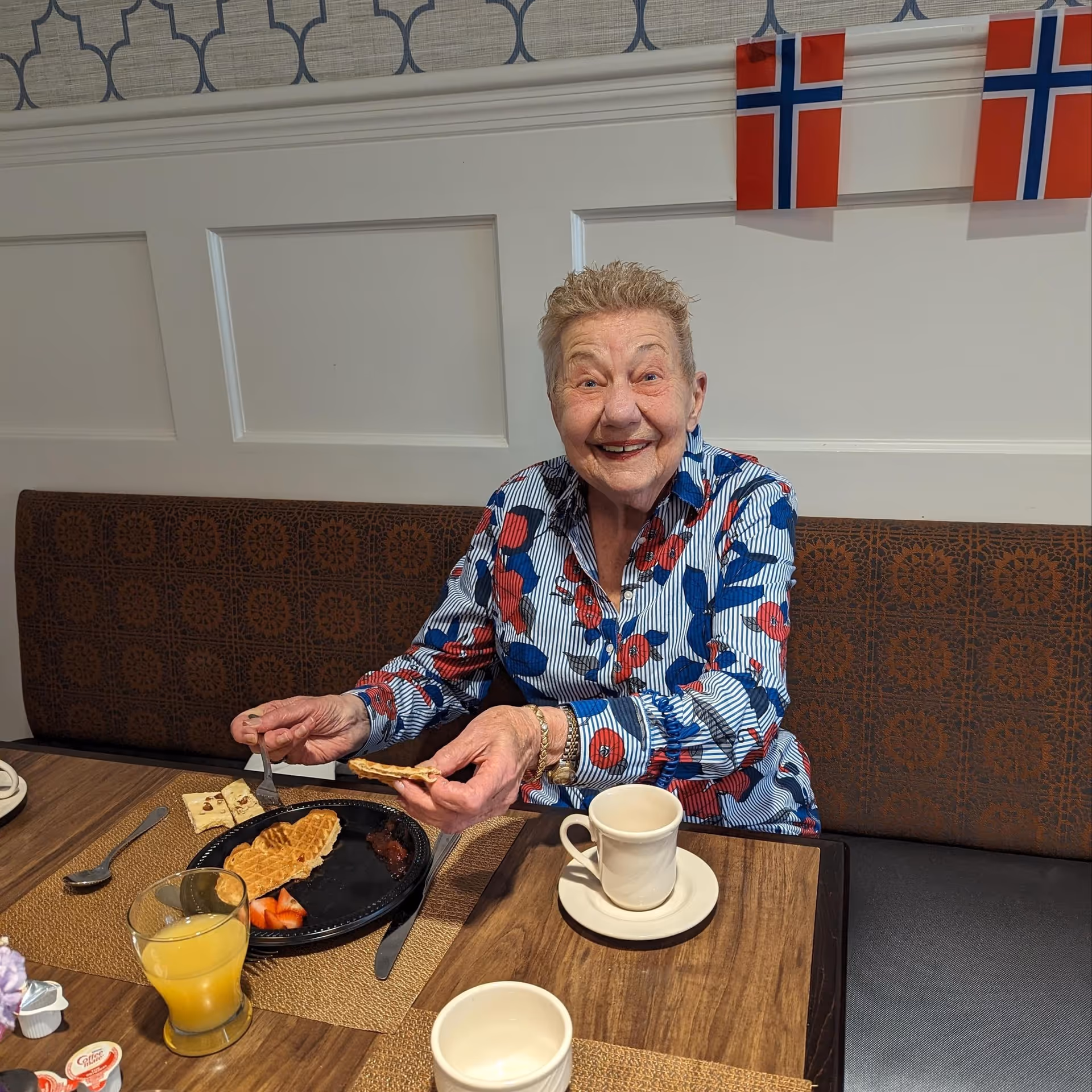 An elderly woman smiling at a dining table, holding a piece of waffle with a plate, cup, and glass of orange juice in front of her.
