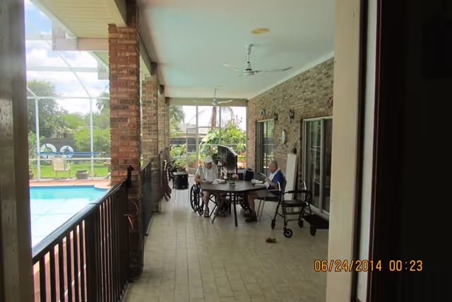 Covered outdoor patio area with tiled floor and brick walls, featuring a round table where two elderly individuals are seated. One person is in a wheelchair and the other is using a walker nearby. The patio overlooks a swimming pool and garden area with greenery and trees in the background.