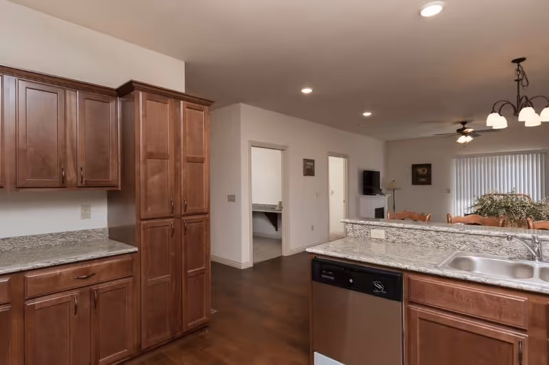 Interior view of a kitchen with wooden cabinets, granite countertops, a stainless steel dishwasher, and a double sink. The kitchen opens into a living area with a dining table, chairs, a ceiling fan, and a window with vertical blinds.