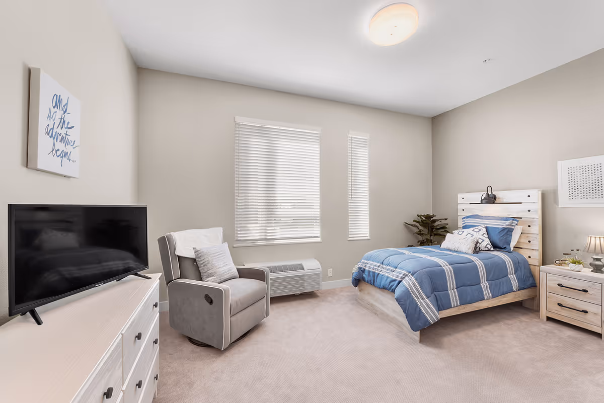 A bright and clean bedroom with beige walls and carpet. The room features a single bed with a blue and white striped comforter, a wooden headboard with a mounted reading light, a wooden nightstand with a lamp and decor, a gray upholstered armchair with a pillow and throw, a white dresser with a flat-screen TV on top, and two windows with white blinds letting in natural light.