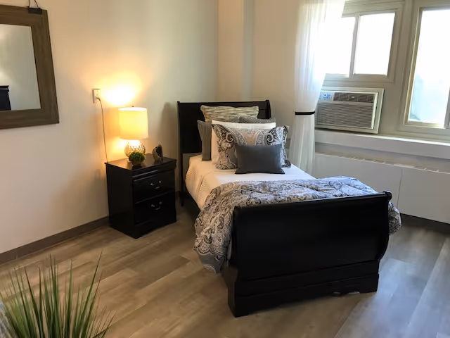 Small furnished bedroom with a single dark wood bed, nightstand and lamp, window air conditioner, mirror, and wood-look flooring.