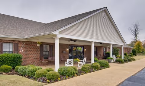 Exterior view of a single-story brick senior living facility with a covered porch supported by white columns. The porch has outdoor seating with chairs and hanging plants. Well-maintained shrubs and flower beds line the walkway leading to the entrance.