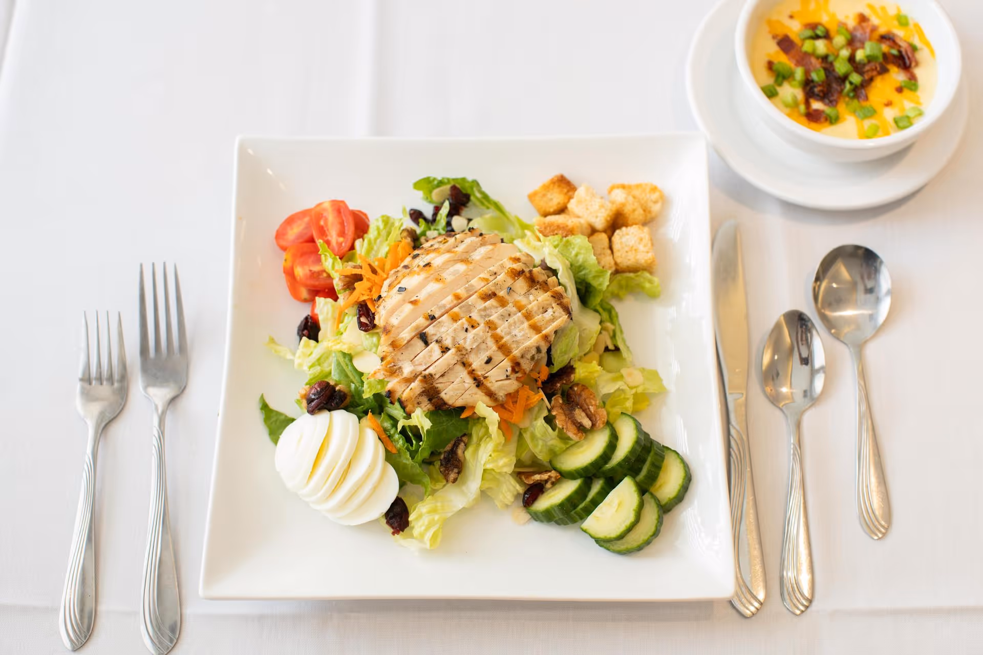 A plated meal consisting of a grilled chicken breast on a bed of mixed salad greens with sliced cucumbers, cherry tomatoes, shredded carrots, croutons, dried cranberries, and a sliced hard-boiled egg. To the right of the plate is a bowl of soup garnished with chopped green onions and bacon bits. The meal is set on a white tablecloth with silverware arranged on both sides of the plate.